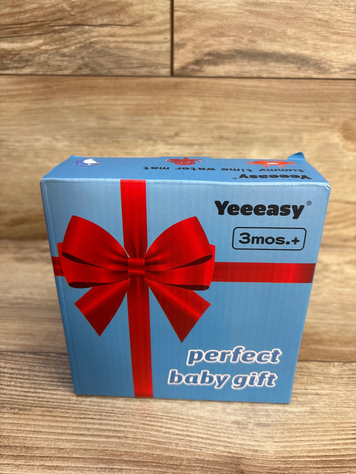 Blue box with red bow, labeled 'perfect baby gift' for ages 3 months and up, on wood surface.