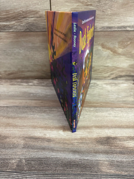 Side view of Dusk Explorers hardcover book showcasing its colorful illustrated cover and spine on a wood-grain surface