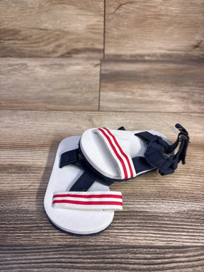 Front view of Child Of Mine baby sandal showing white footbed, navy straps and red-and-white striped straps on wood floor