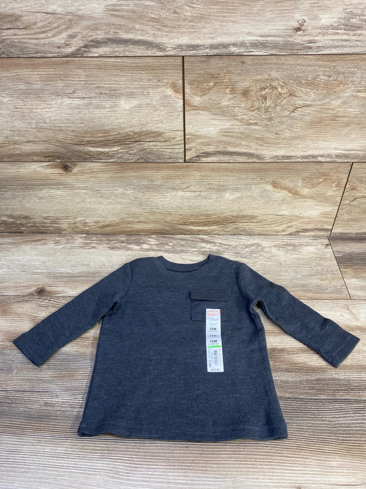 Flat lay of Jumping Beans long sleeve baby shirt showcasing ribbed knit texture, chest pocket, and visible size tag on light wood background