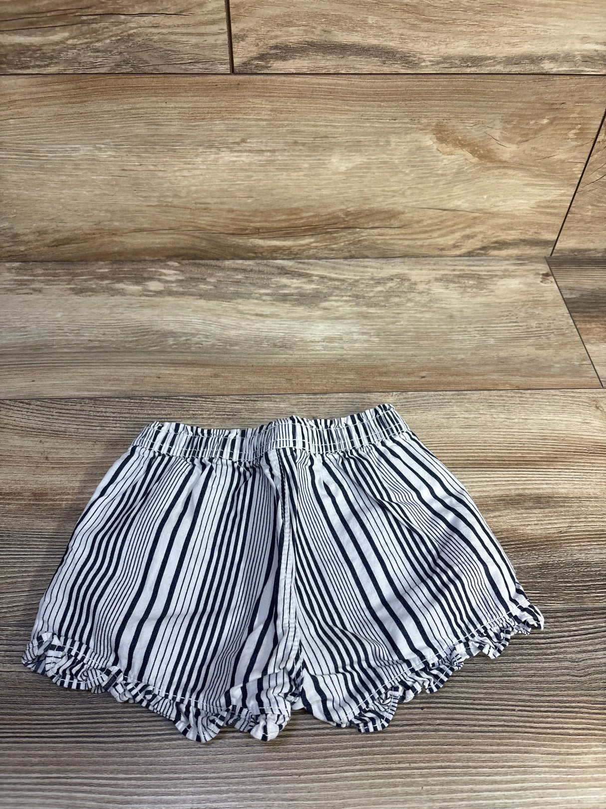 Top-down view of Baby Gap white and navy striped toddler shorts showcasing elastic pull-on waistband and scalloped ruffle hem on wood background