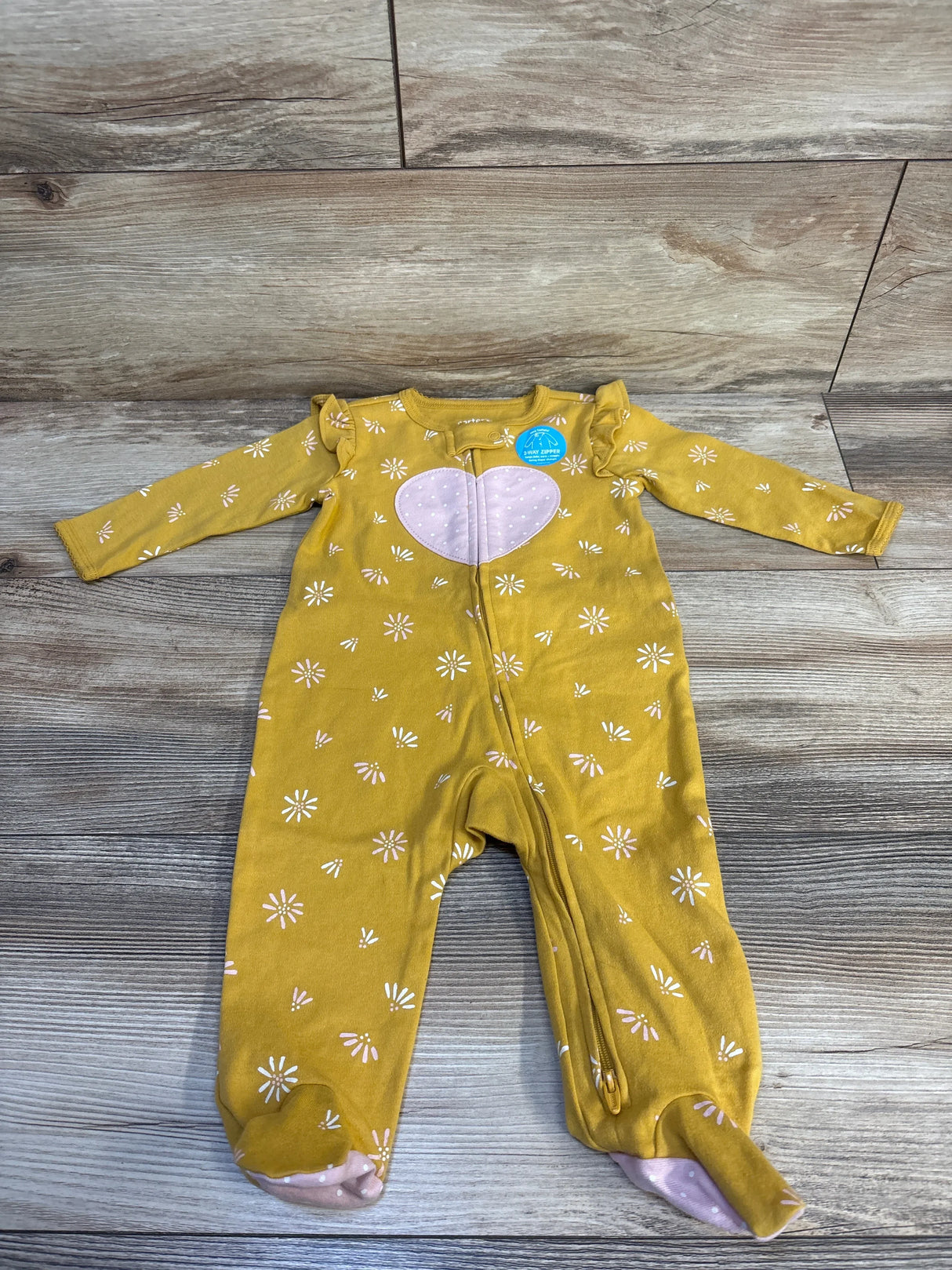 Mustard yellow baby footed onesie with white daisies, pink heart, and 2-way zipper, laid on wood floor