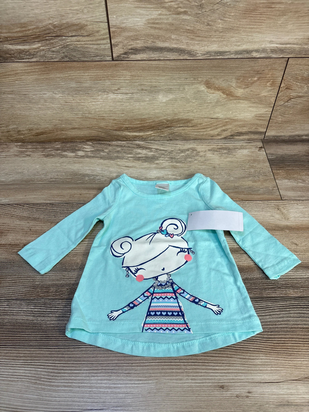 Mint green long sleeve toddler shirt with girl cartoon graphic, laid on wood floor