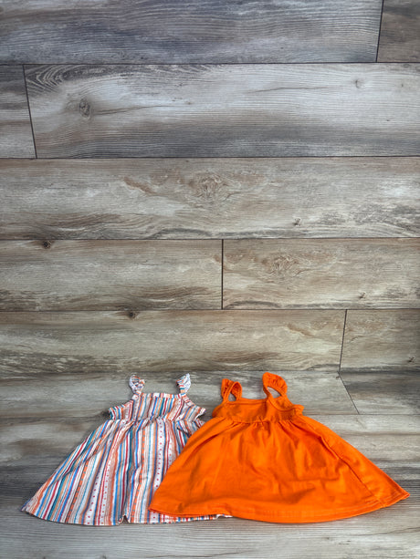 Flat lay of two baby sleeveless dresses showing a solid bright orange dress and a multicolor striped dress on wood background