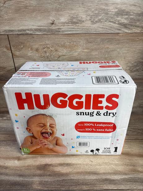 Front of Huggies Snug and Dry size 1 box, 108 diapers, baby photo and leakproof claim on wood surface