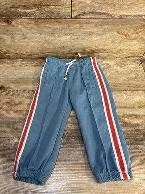 Blue kids joggers with red and white side stripes, elastic waist and cuffs, laid on wood floor