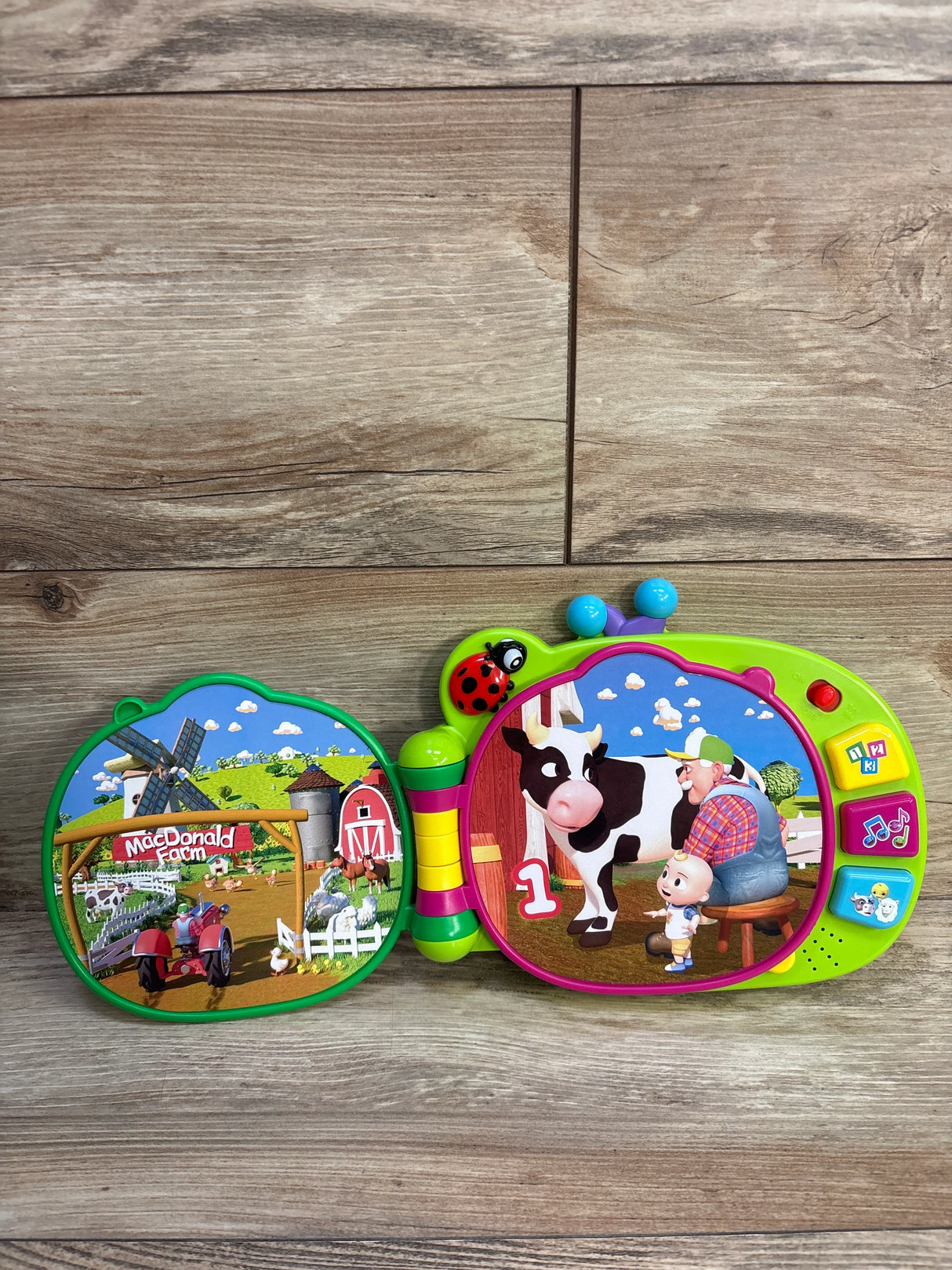 Front view of Cocomelon MacDonald Farm learning book showing cow and farmer, buttons and ladybug handle on wood floor