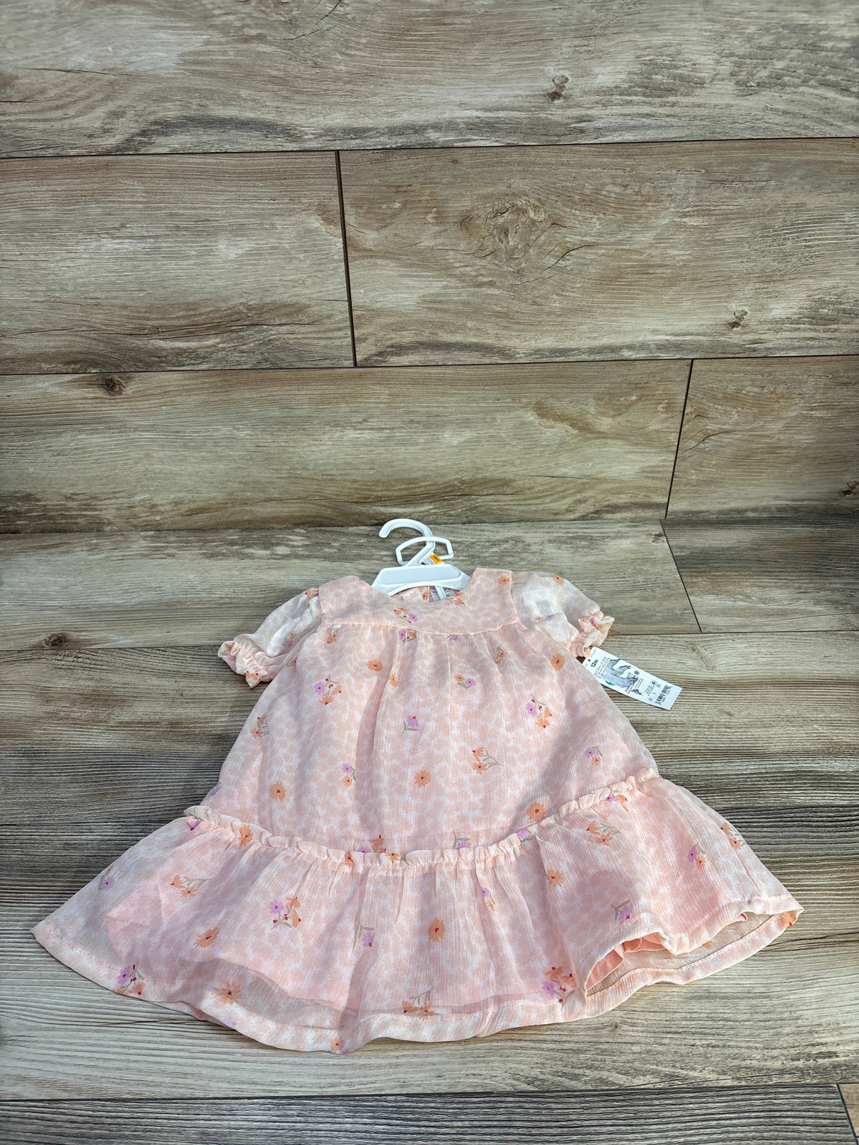 Flat-lay of pink floral baby dress (12M) showcasing ruffled hem and puff sleeves with wood plank background