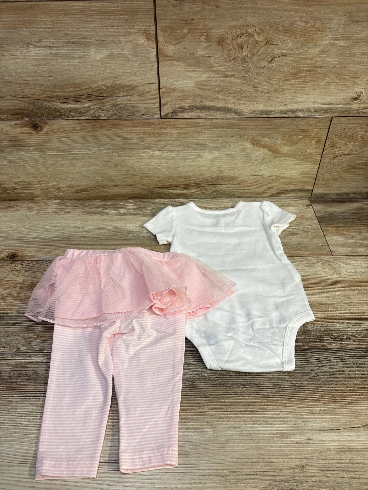 Top-down view of Carter’s two-piece bodysuit and skeggings set showcasing a white bodysuit and pink skirted leggings on a wood-grain background