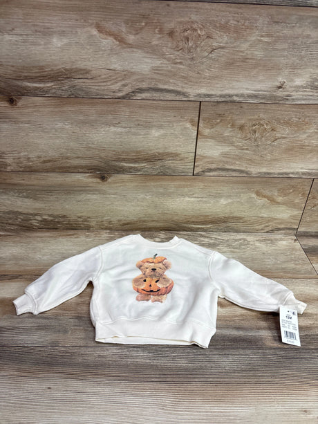 Front view of the ivory Grayson Mini Pumpkin Bear sweatshirt with long sleeves and pumpkin bear graphic on a wood floor