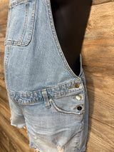 Side close-up of Isabel maternity denim shortalls showcasing side button closures, front bib pocket, and frayed hem on wood background