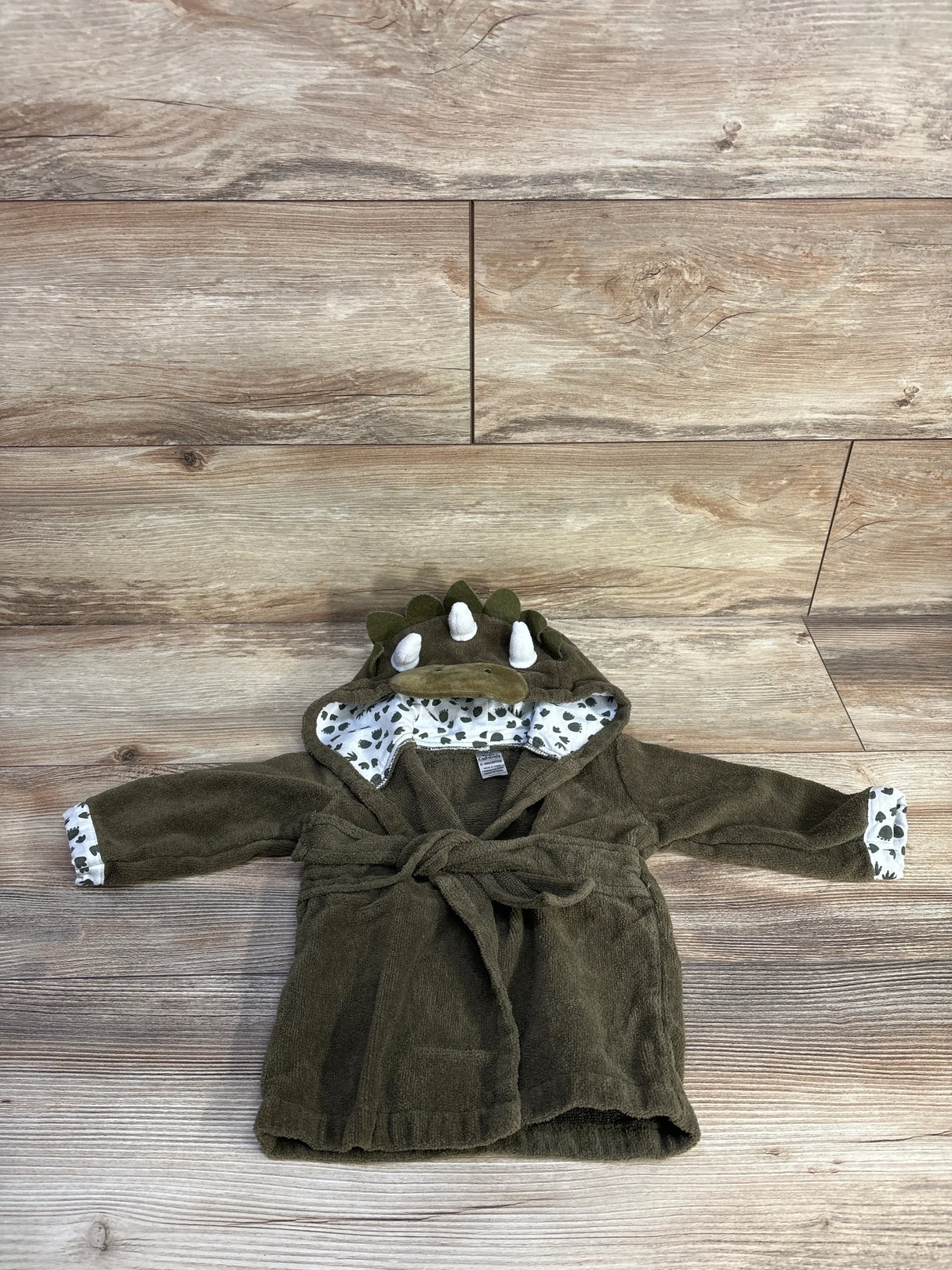 Flat lay of green dinosaur baby bath robe showing hood spikes, printed leaf lining, and tied waist belt on light wood background