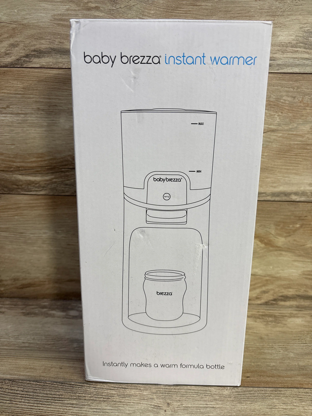 Front view of Baby Brezza Instant Bottle Warmer box showing line illustration of the warmer and the headline 'Instantly makes a warm formula bottle' on a wood backdrop