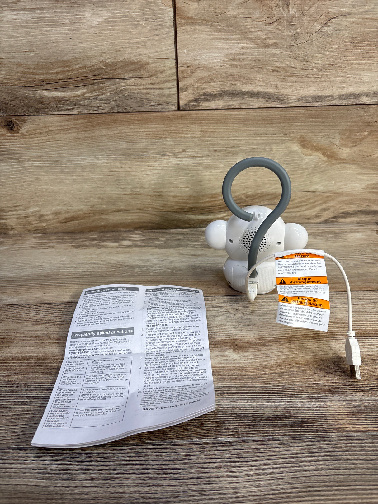 Rear view of white VTech Myla monkey soother with gray hanging loop, USB cable, and manual on wood background