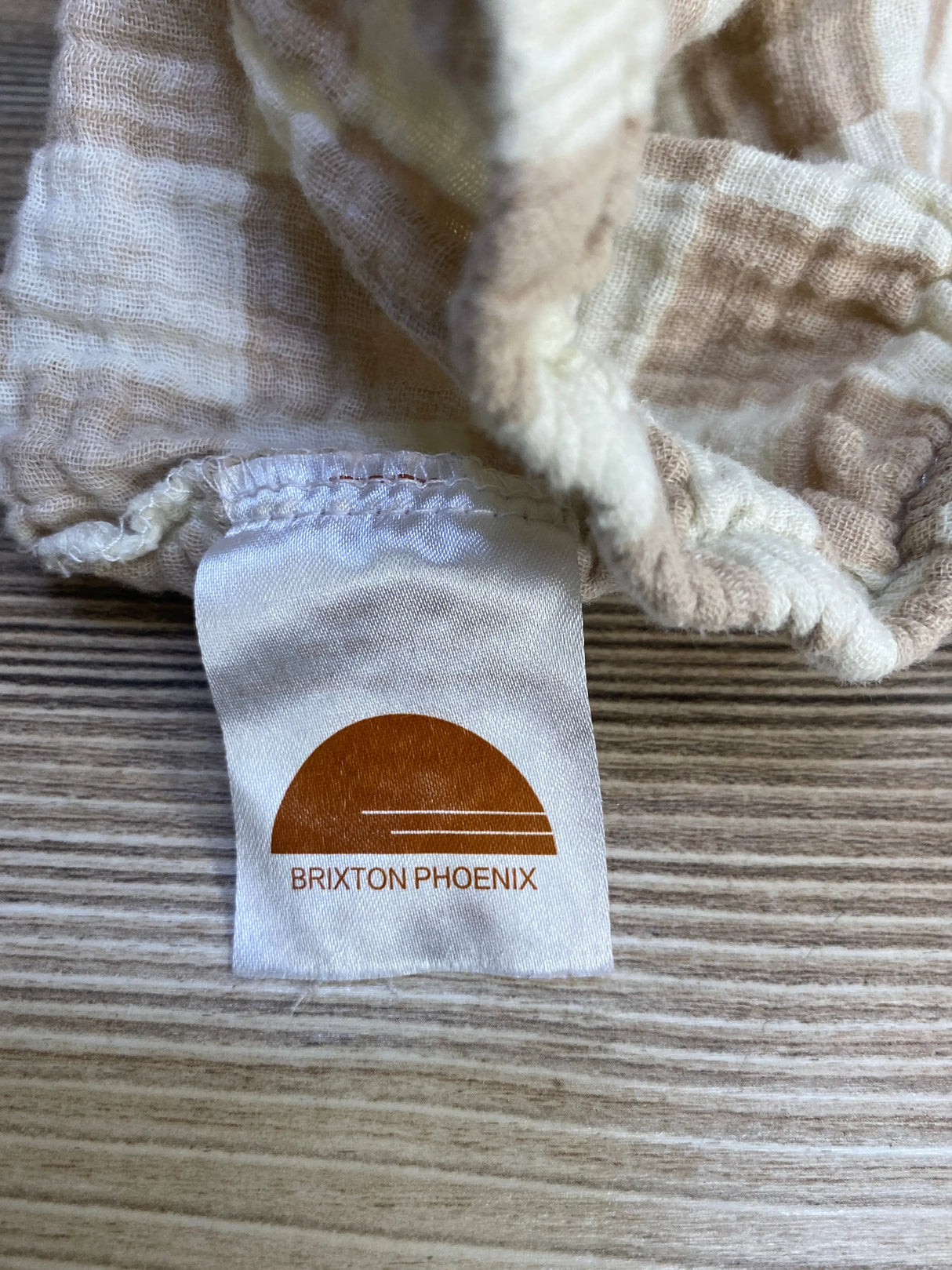 Close-up of Brixton Phoenix muslin bassinet sheet showing brand label and beige check fabric on wood background