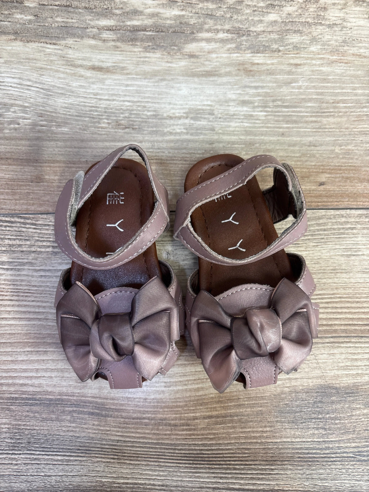 Top-down view of mauve baby sandals showcasing satin bows, ankle Velcro straps and cushioned insoles on light wood floor