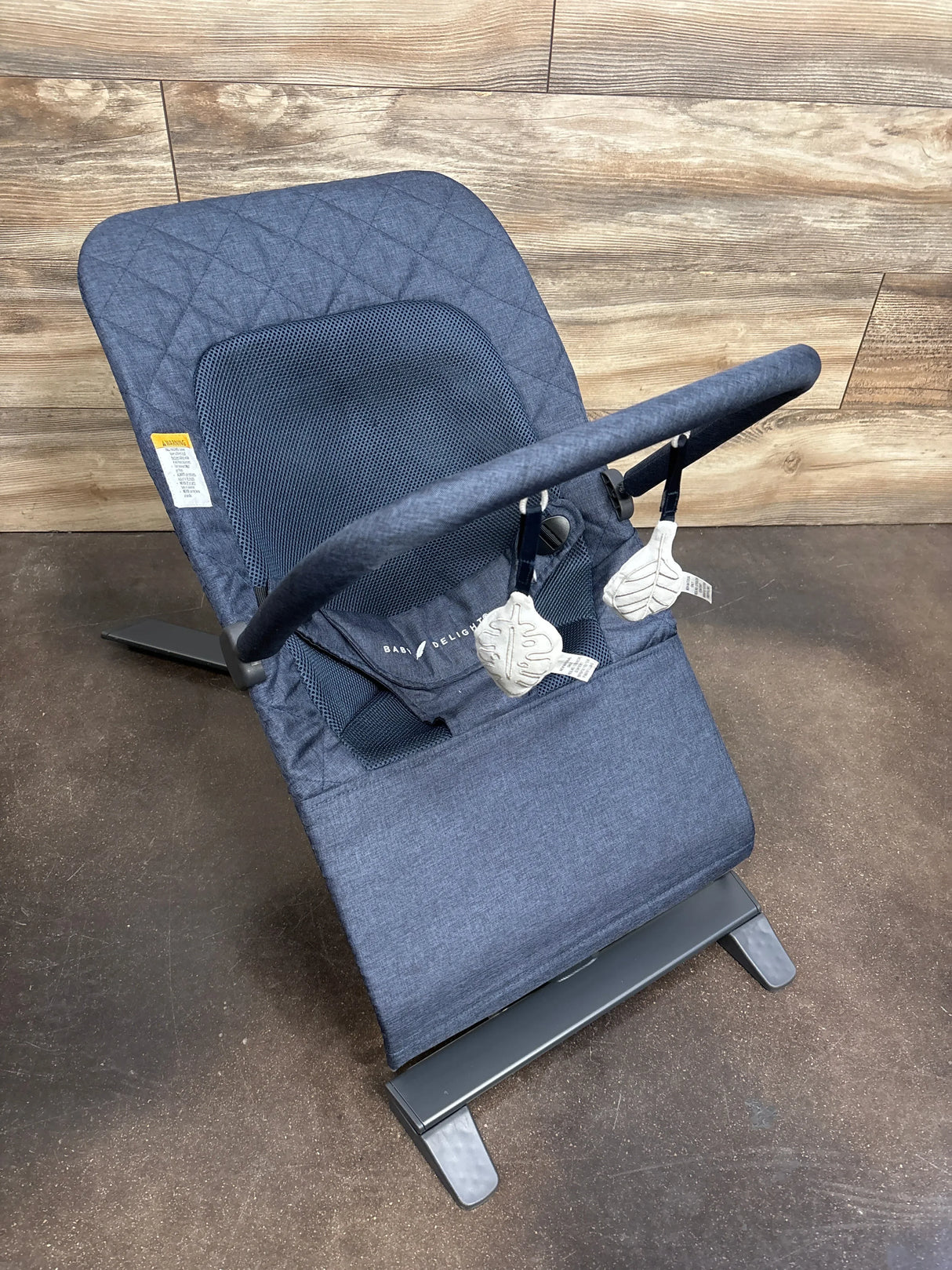 Three-quarter view of Baby Delight Alpine Deluxe bouncer in quilted indigo showcasing mesh seat and toy bar on wood plank floor