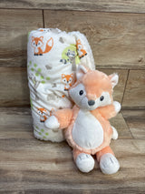 Angled front view of plush baby blanket with fox and raccoon print and matching peach fox plush on wood floor