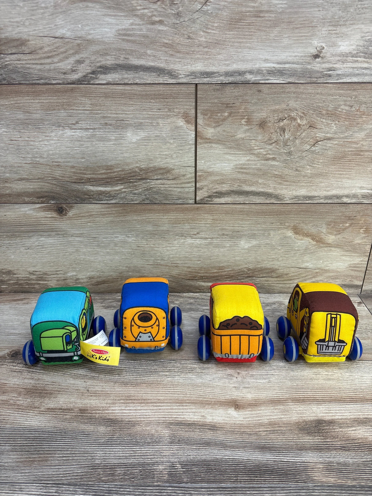 Front view of four K's Kids plush pull-back construction trucks in green, blue and yellow on wood background