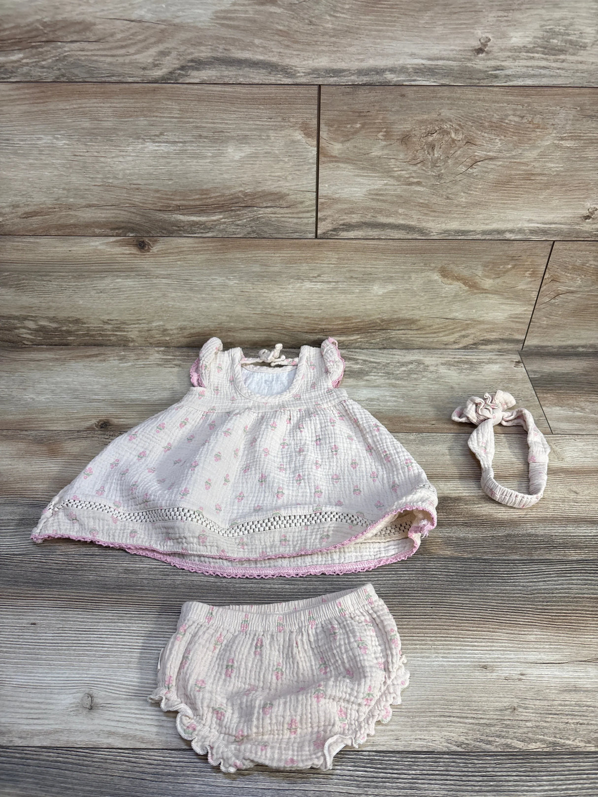 Flat lay of beige 3-piece embroidered gauze baby dress set size 12m with dress, bloomers and headband on wood background