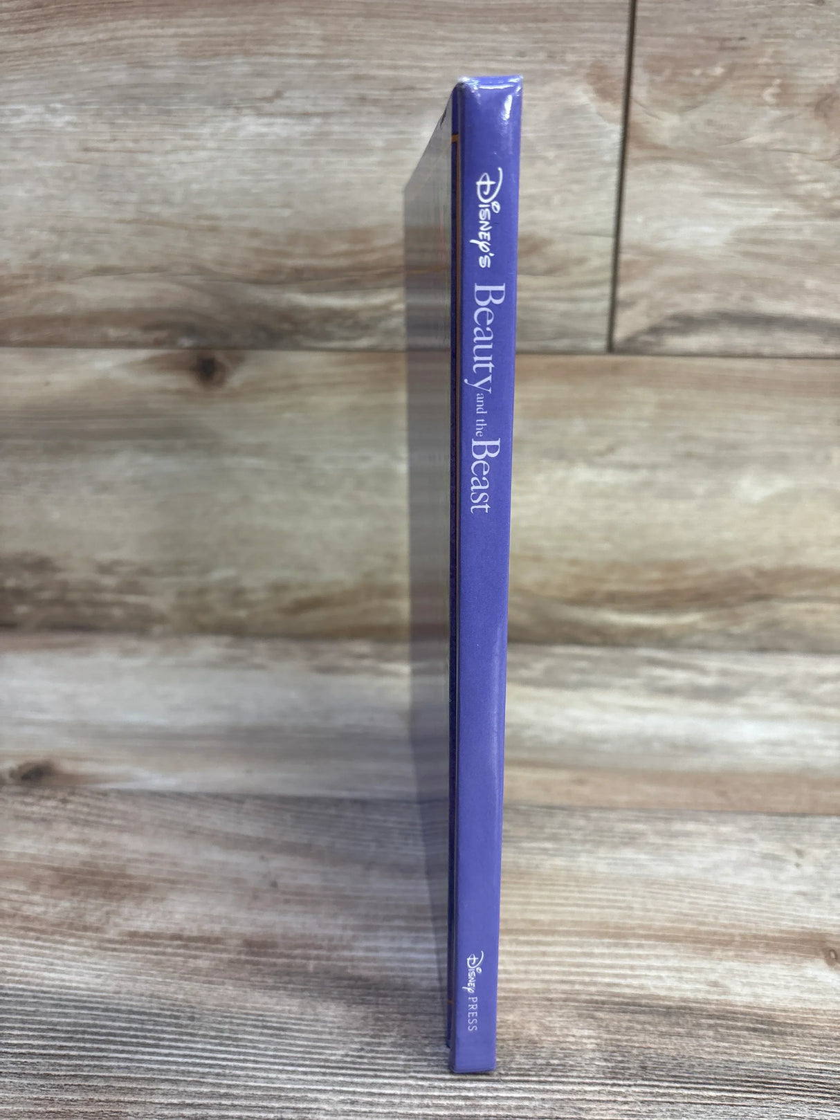 Side view of Disney Beauty and the Beast hardcover showing purple spine with title text and shrink-wrap on wood background