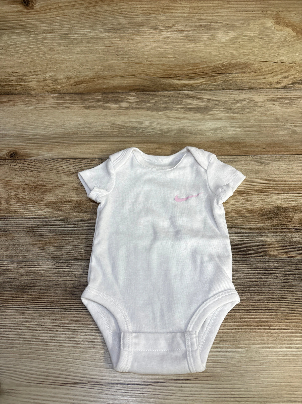 Flat lay of white Nike baby bodysuit showcasing small pink Swoosh logo on wood floor background