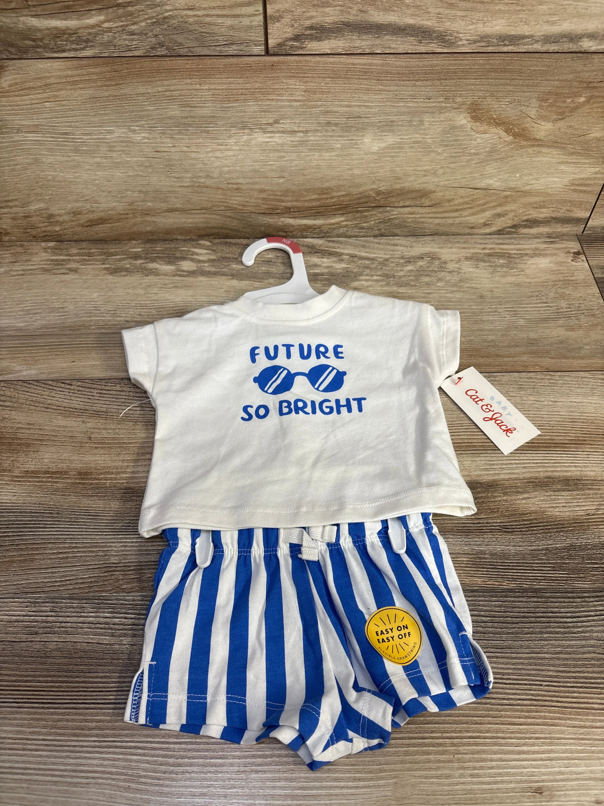 Front view of Cat & Jack newborn two-piece: white tee reading FUTURE SO BRIGHT and blue-white striped shorts on wood background