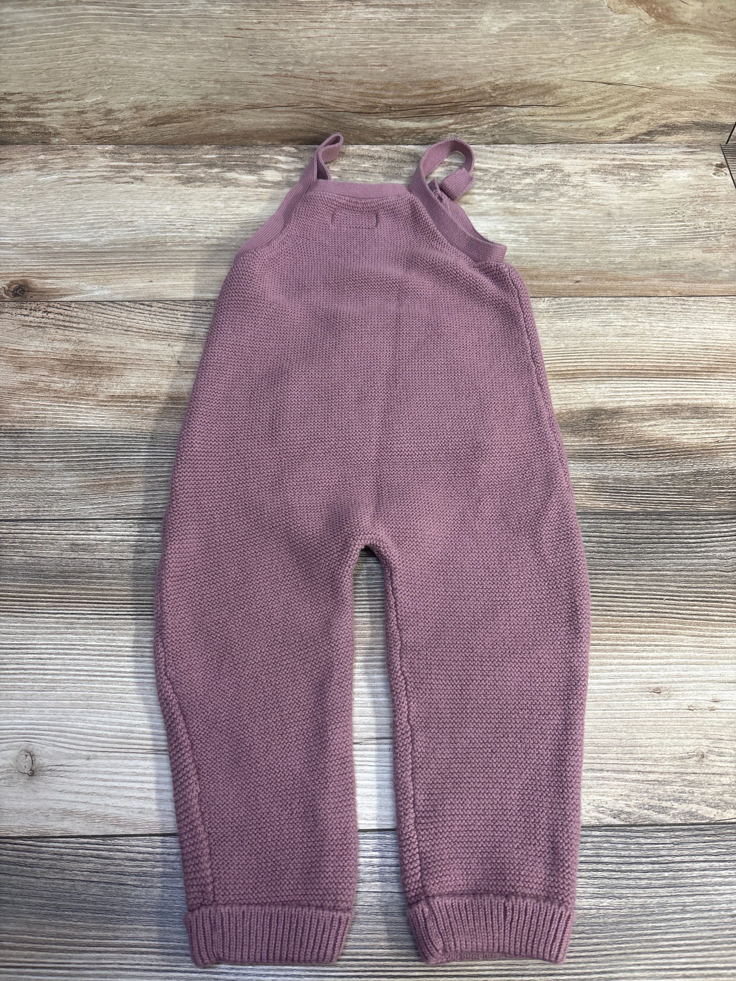 Top view of Little Planet knit overalls in mauve showcasing shoulder straps and ribbed cuffs with a wood grain background