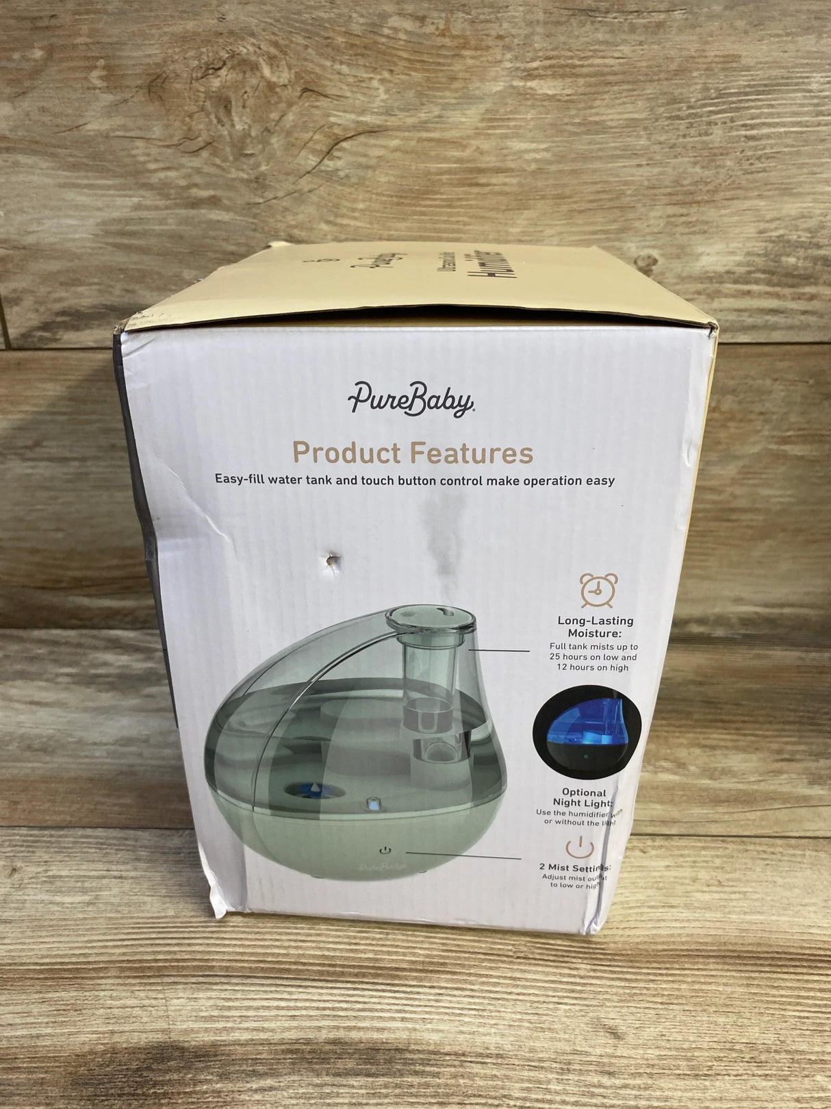 Front view of boxed PureBaby ultrasonic cool mist humidifier showcasing clear water tank and product features text, on wooden surface