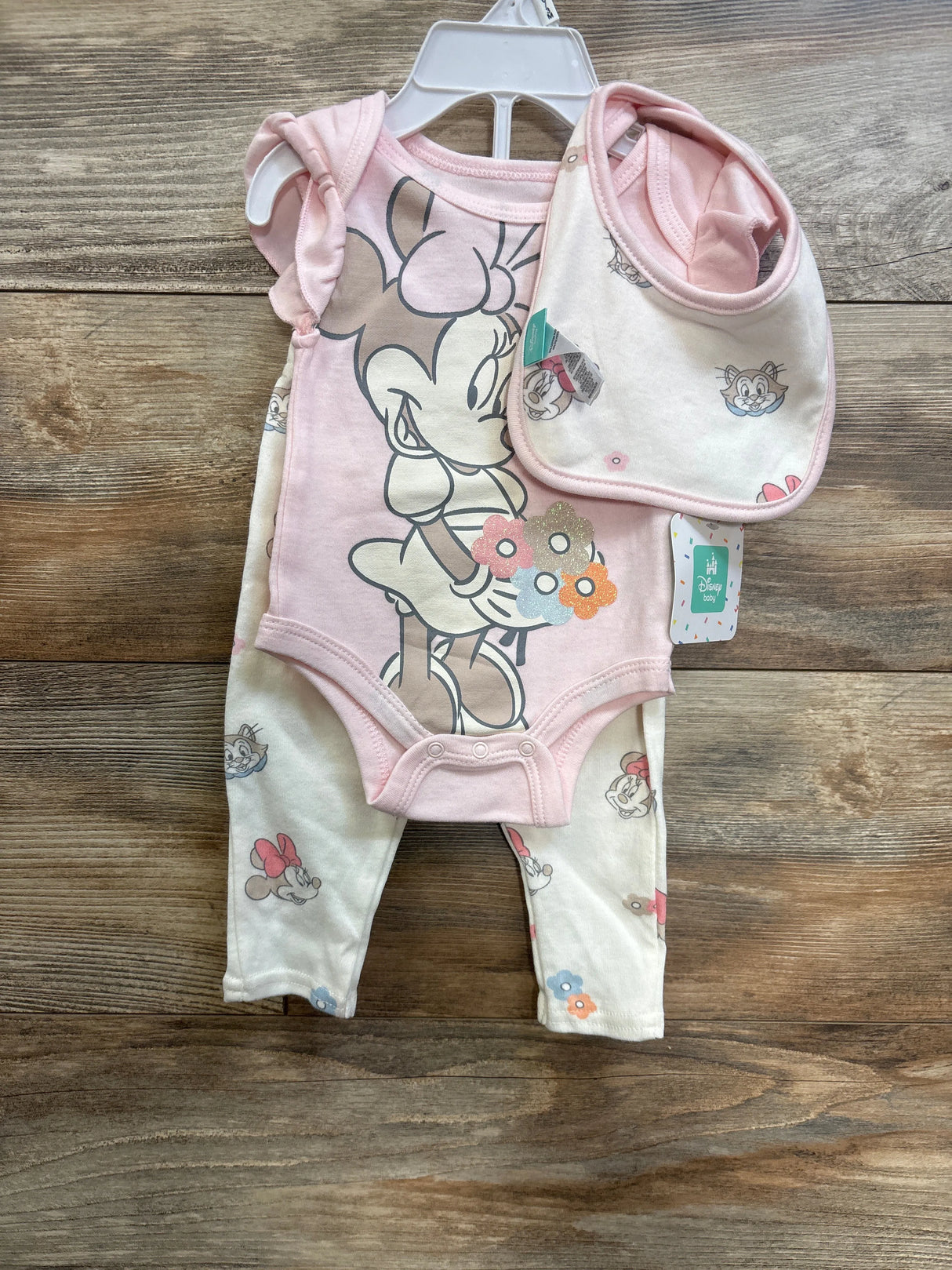 Front view of pink Minnie Mouse 3-piece baby set: bodysuit with large Minnie print, matching pants and bib on wood background