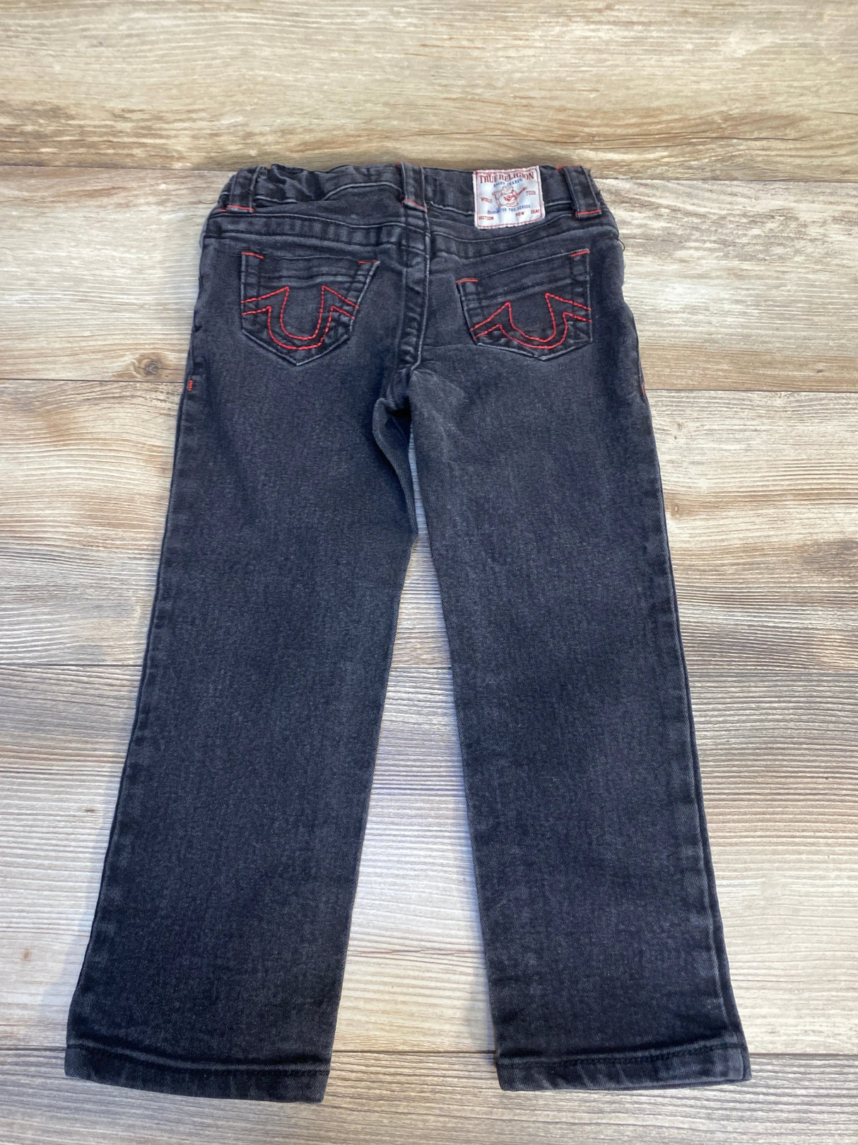 Black kids jeans with red stitching, back pockets, and logo patch on wood background