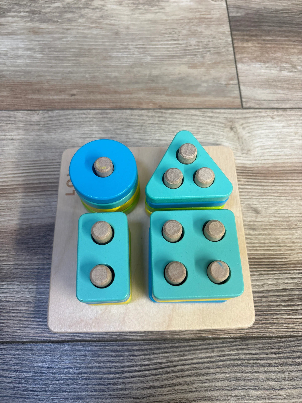 Top view of Lovevery Sort and Stack Peg Puzzle showcasing blue circle teal triangle rectangle and square pegged shapes on wood plank floor