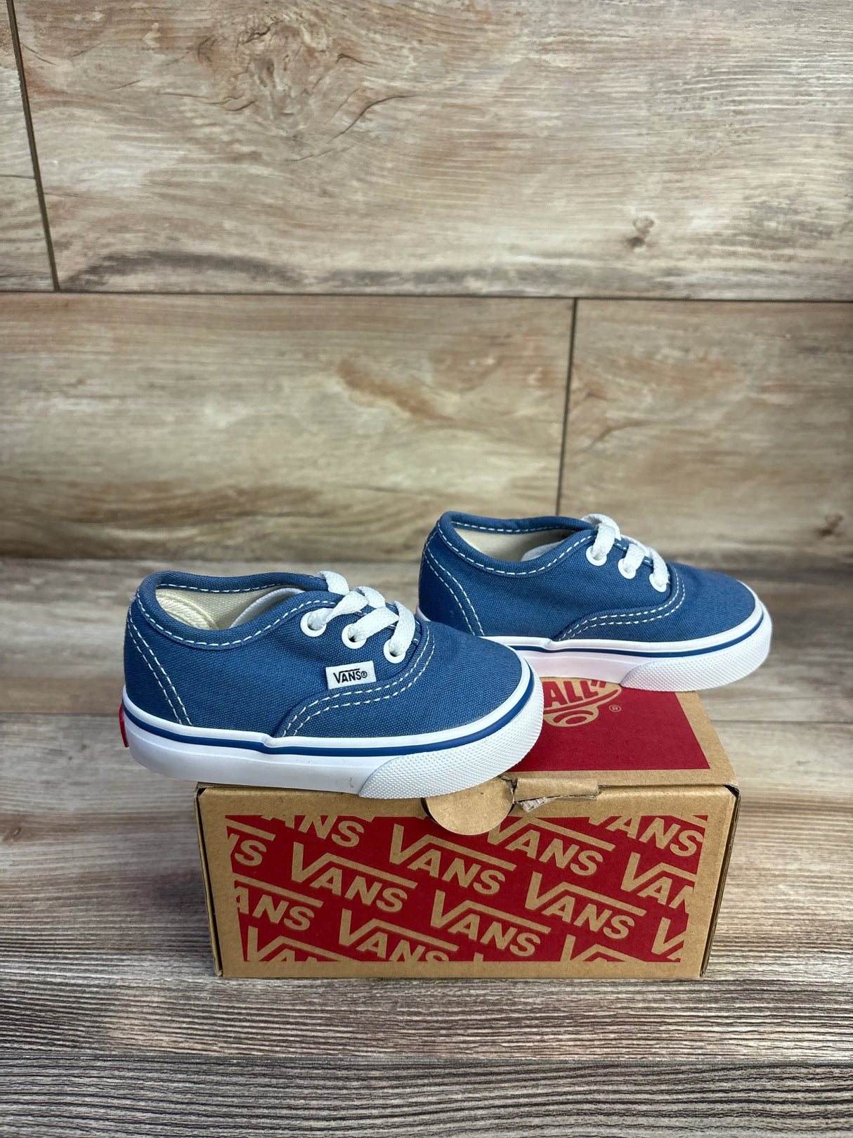 Side view of navy Vans Authentic low-top toddler sneaker showing white laces, white sole stripe, and Vans tag on a Vans box with wood background