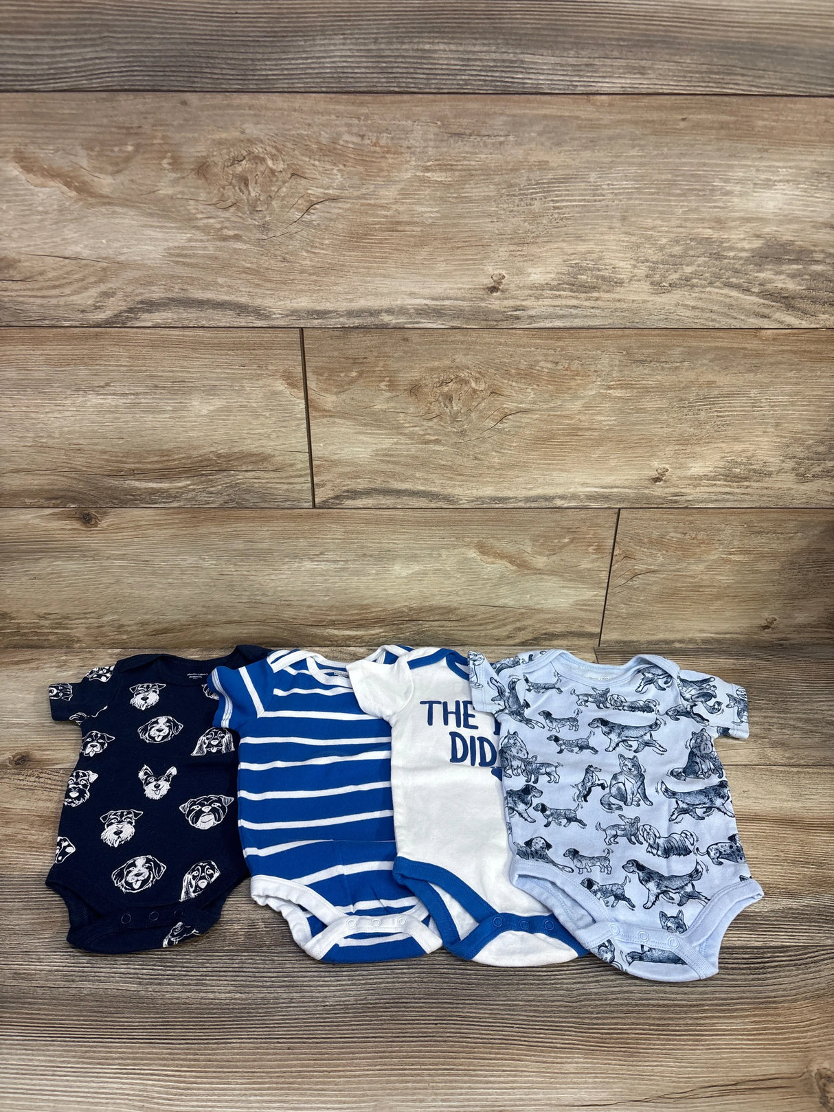 Front view of 4 pack light blue 6 month baby bodysuits showing dog print, striped, and text designs on wood background