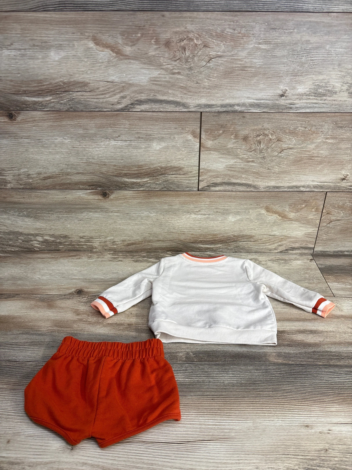 White long-sleeve baby shirt with orange trim and matching orange baby shorts on wood floor