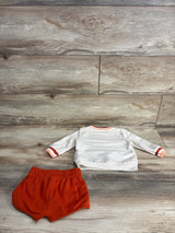 White long-sleeve baby shirt with orange trim and matching orange baby shorts on wood floor