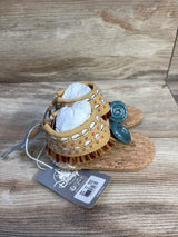 Three-quarter view of toddler Moana sandals with cork soles, tan shell-pattern ankle cuffs, brown fringe, and teal spiral toe accents on wood