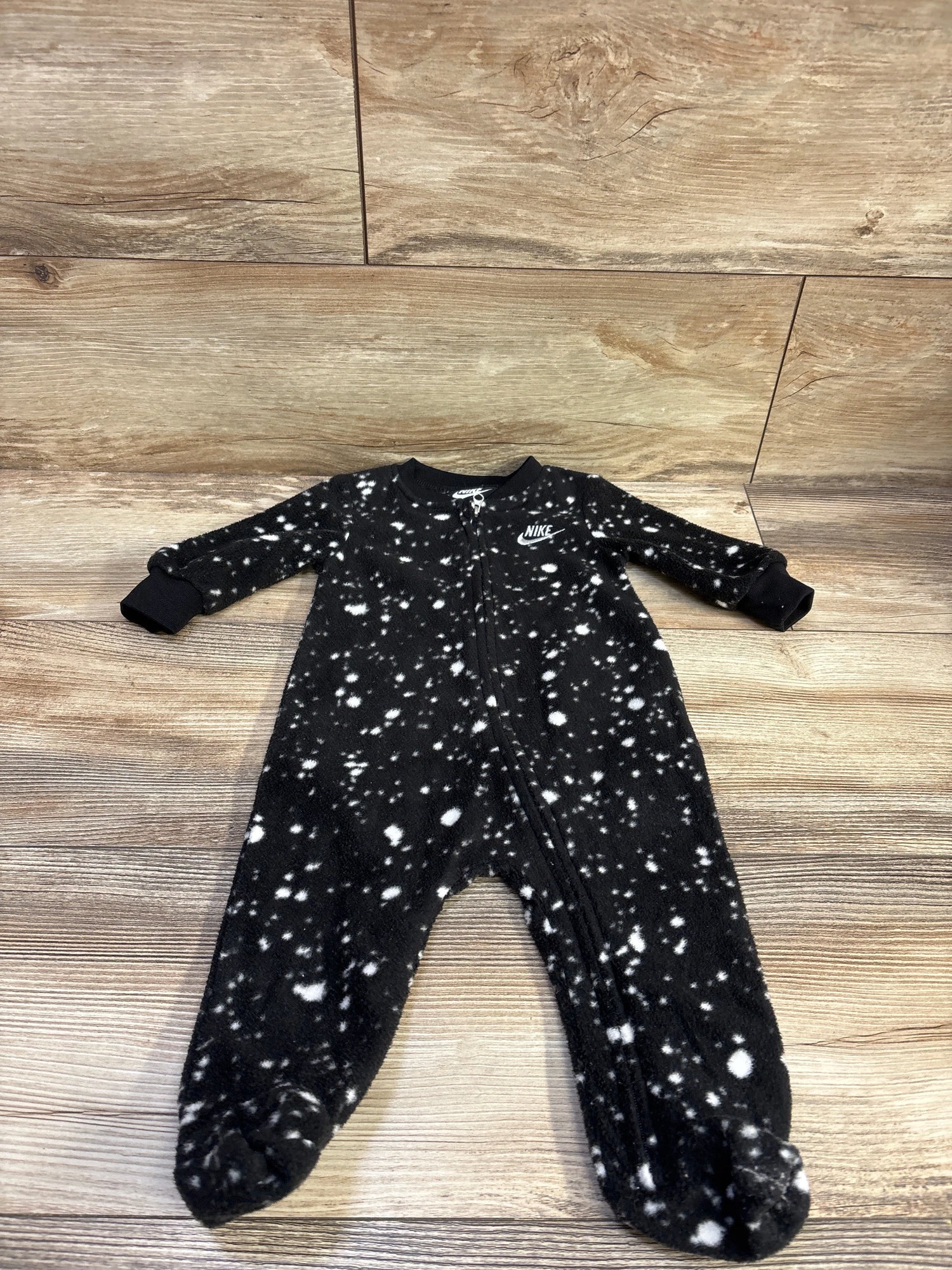 Front view of Nike black blanket sleeper in size 6m showcasing white splatter pattern, front zipper, and covered feet on wood floor