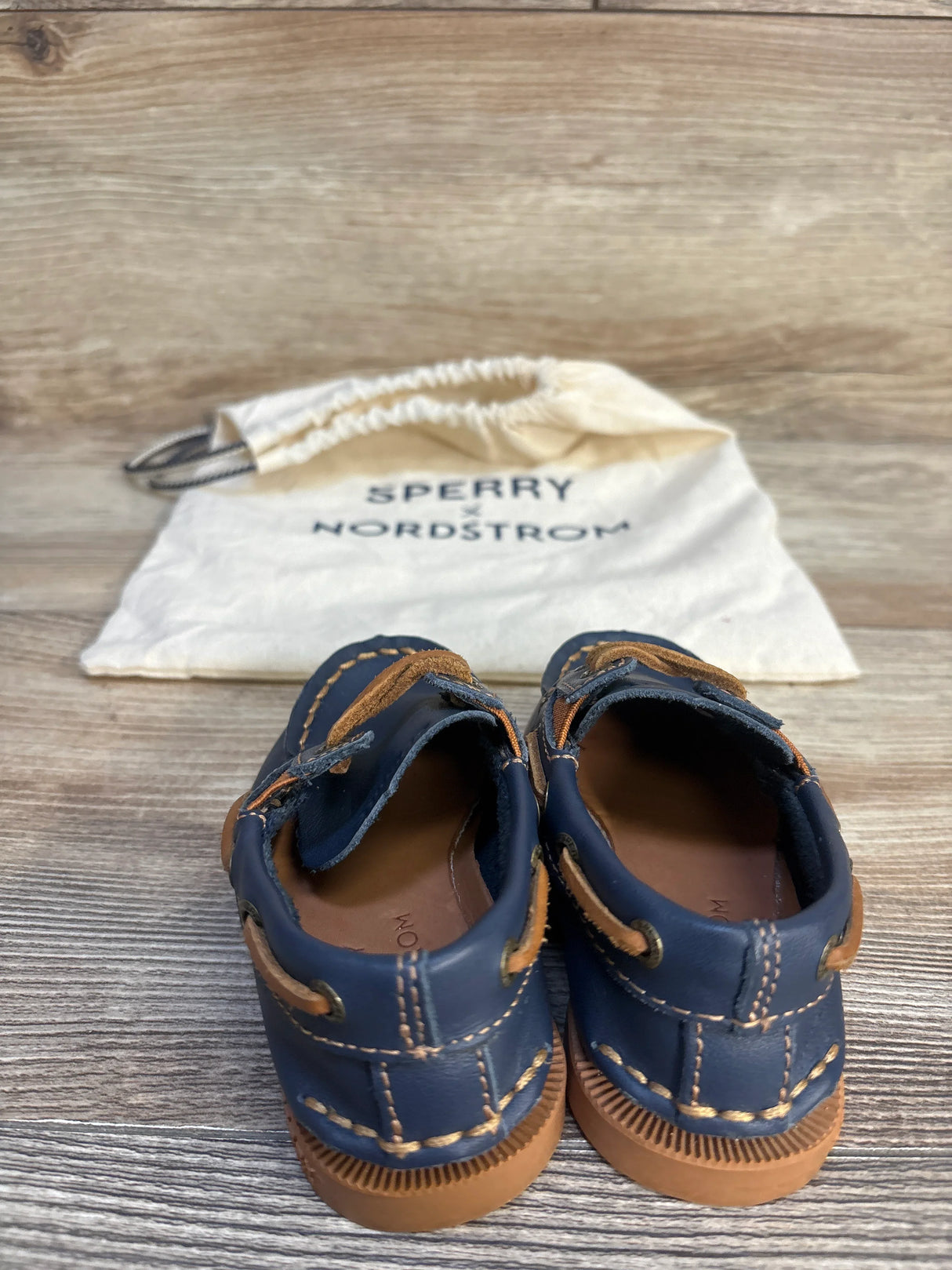 Navy blue and brown toddler boat shoes with stitched details, placed in front of a Sperry Nordstrom drawstring bag on wood background.