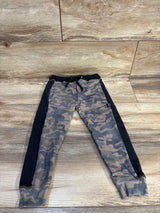Kids camo jogger pants with black side stripes and drawstring, laid flat on wood floor