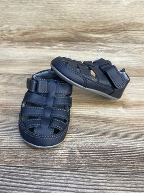 Angled view of navy Harrison fisherman baby sandals showing hook and loop ankle strap and woven upper on wood background
