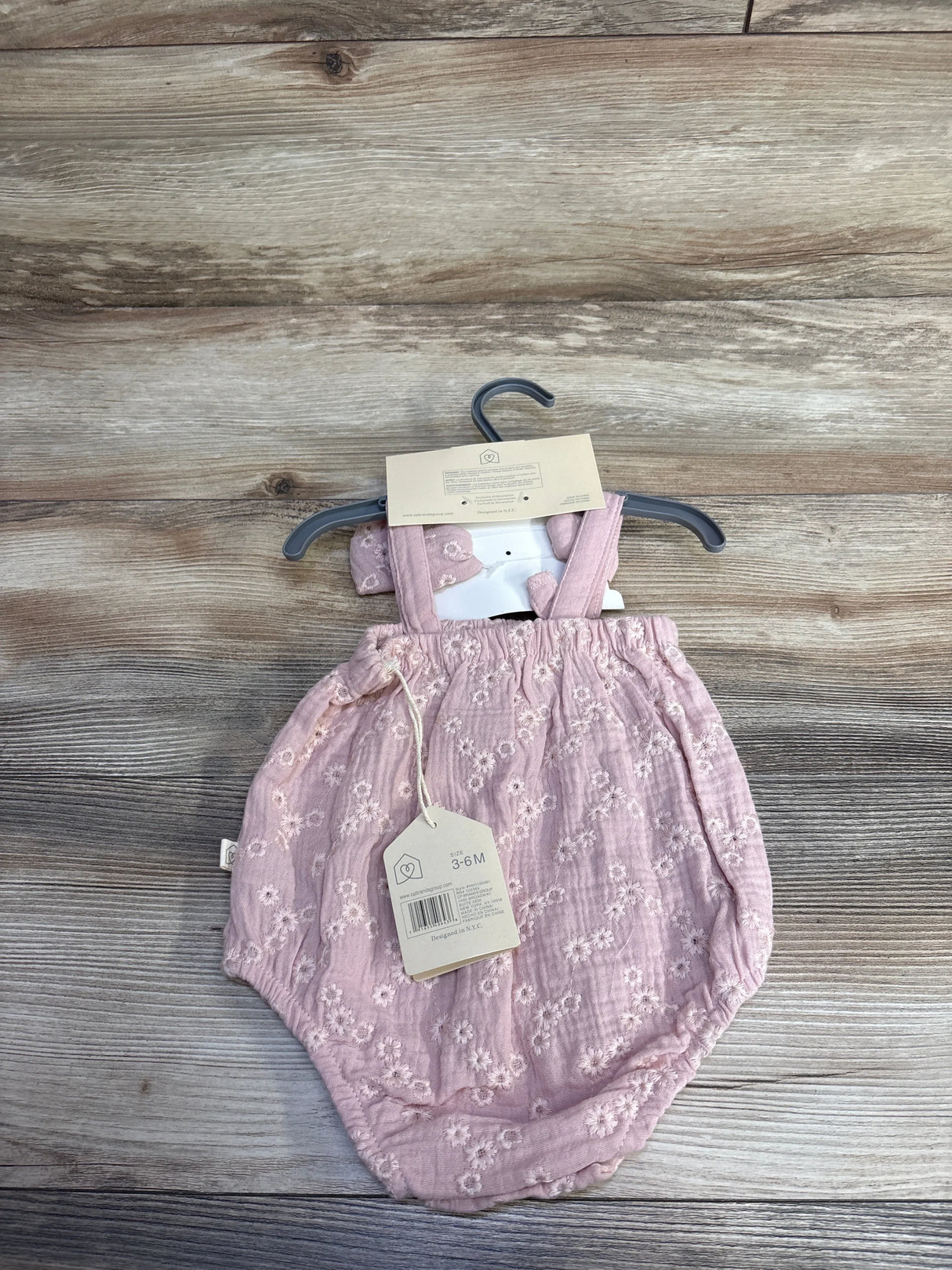 Front view of pink embroidered bubble romper and matching headband on hanger, showing floral embroidery and size 3-6M tag, wood background