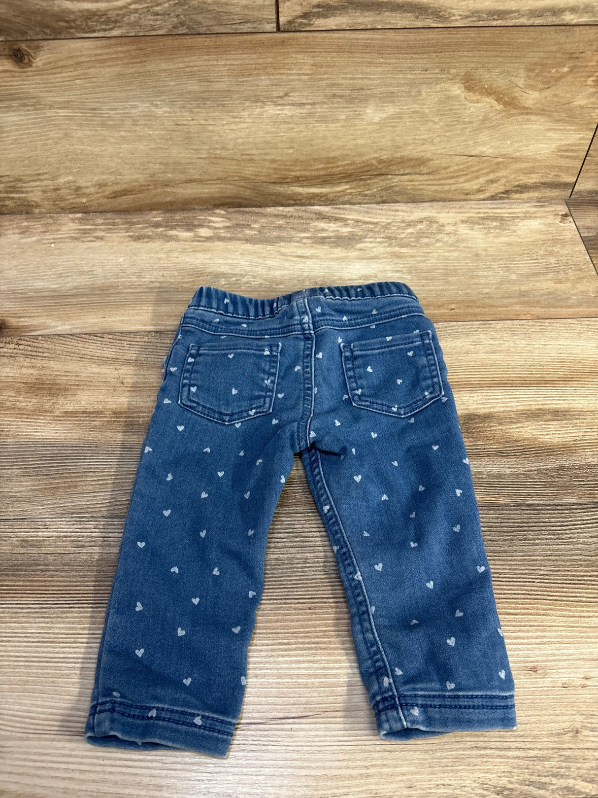 Back view of blue Cat & Jack jeggings with elastic waistband, back patch pockets, and white heart print on wood background