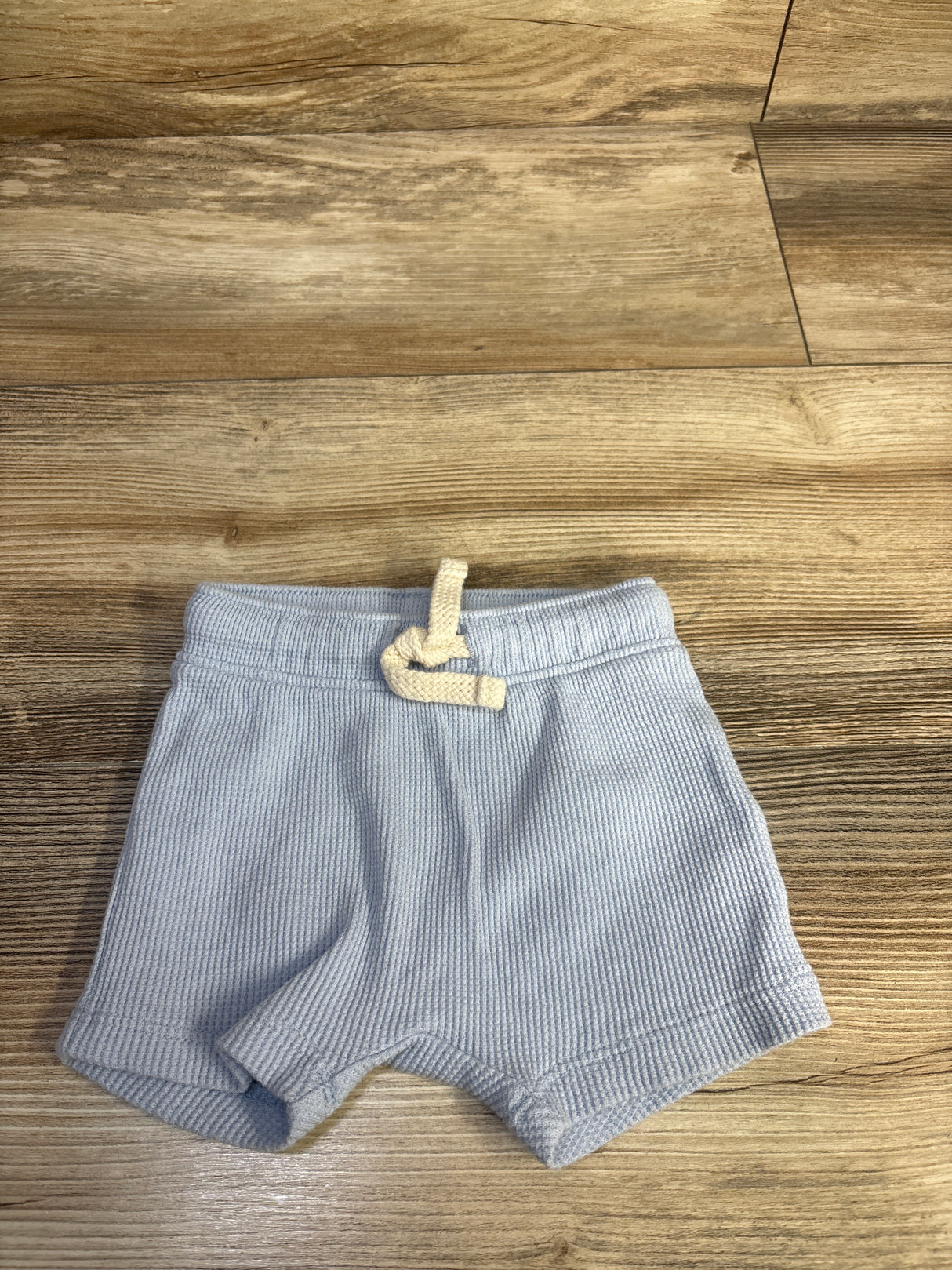 Flat lay of newborn blue waffle-knit pull-on shorts showing elastic waistband with cream drawstring on wood floor