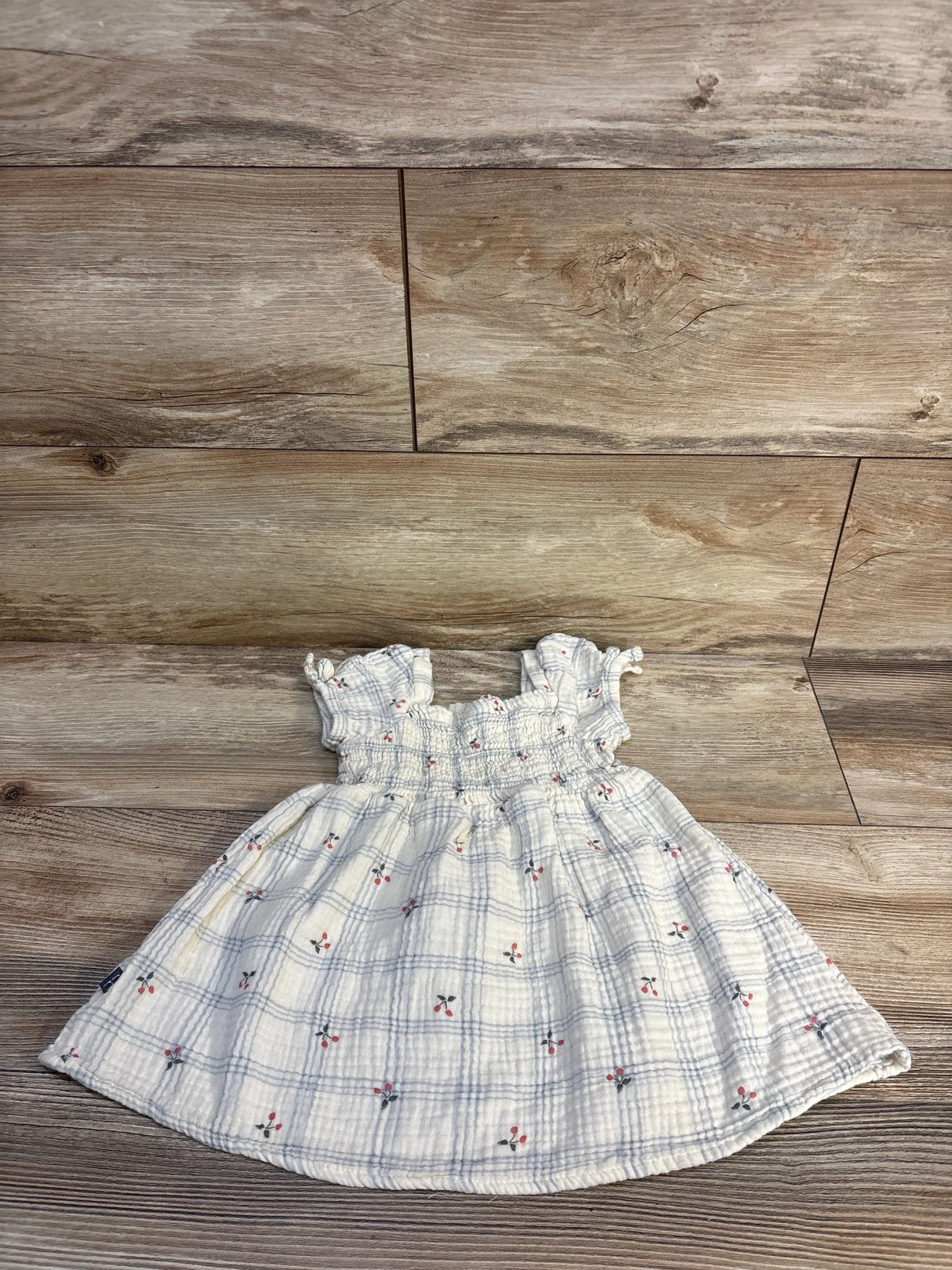 Flat lay of ivory smocked muslin baby dress showing smocked bodice, puff sleeves and small cherry print on wood floor