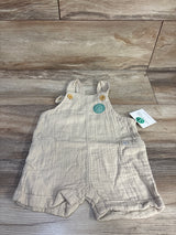 Front flat lay of Little Planet beige organic gauze shortalls (9M) showing wooden-button straps and front pockets on wood floor