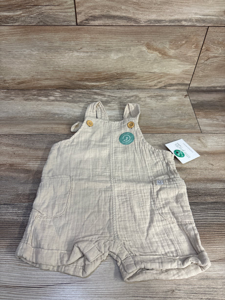 Front flat lay of Little Planet beige organic gauze shortalls (9M) showing wooden-button straps and front pockets on wood floor