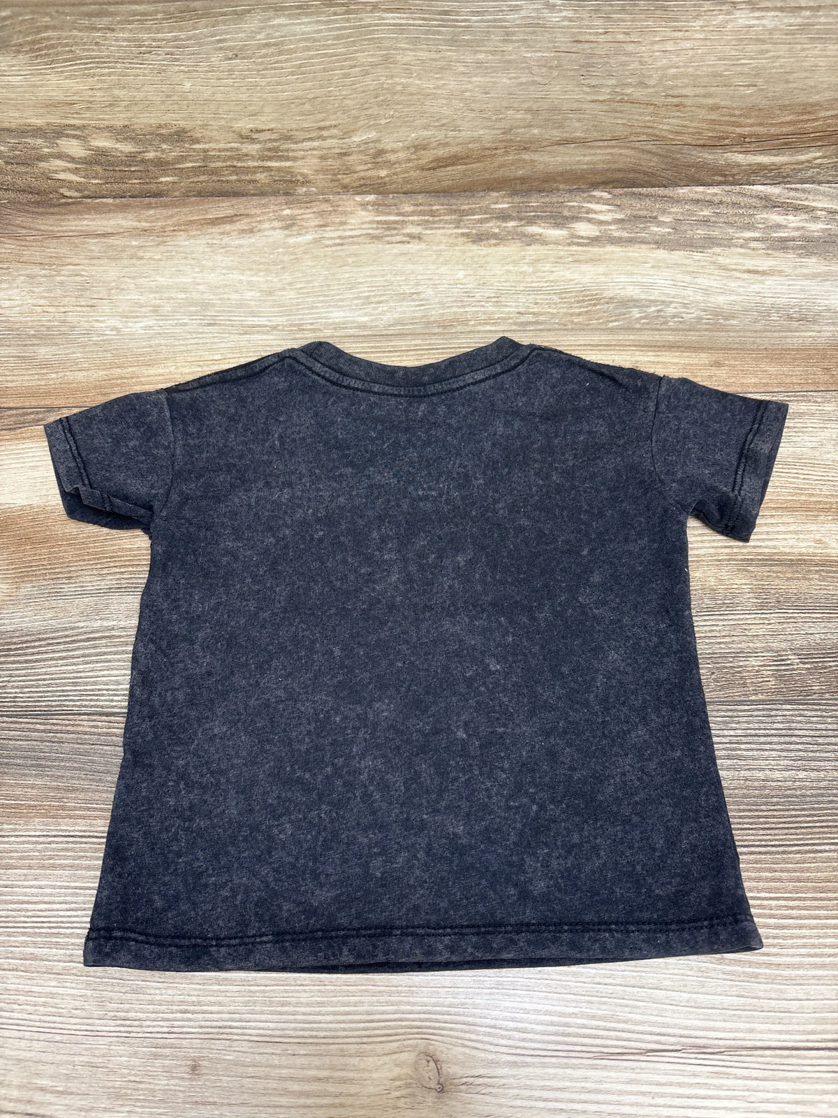 Back view of The Rolling Stones black shirt, size 18 months, showing short sleeves and a plain back on a wood surface