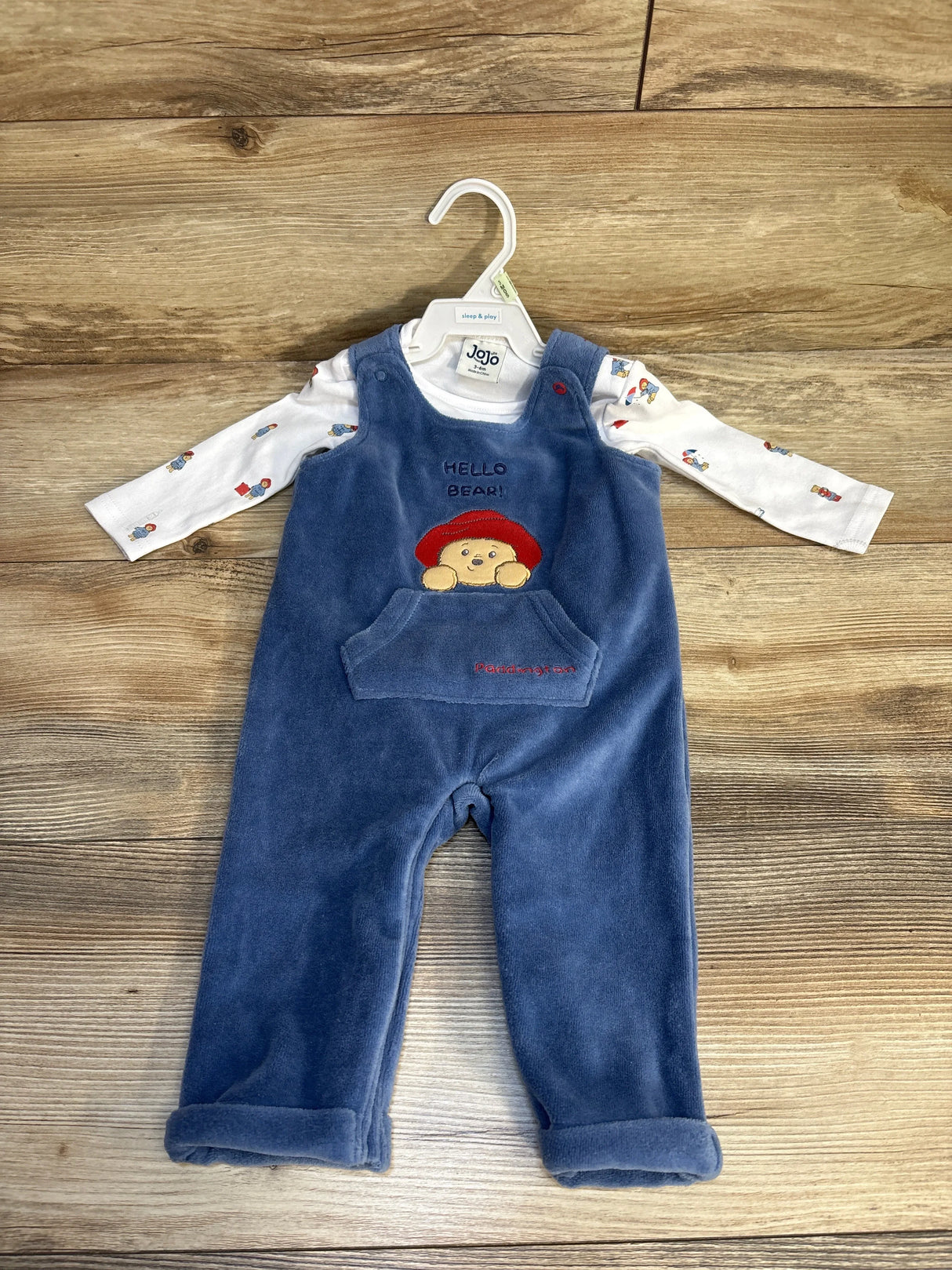 Front view of baby overalls and shirt set showcasing blue velour overalls with Paddington bear motif over white long sleeve on hanger on wood