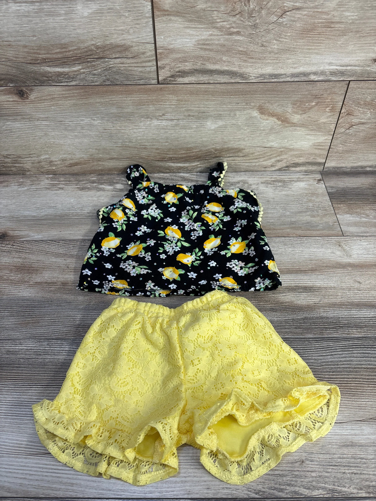 Flat lay of toddler two-piece set: black lemon-print top with gem accents and yellow lace ruffle shorts on wood background